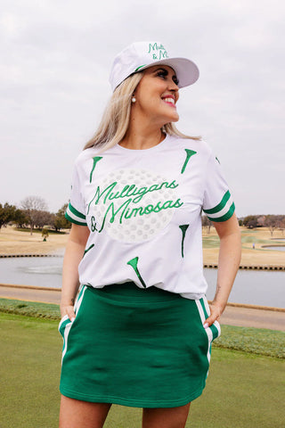 Woman in golf attire with a white Mulligans & Mimosas shirt and green skirt on a golf course.