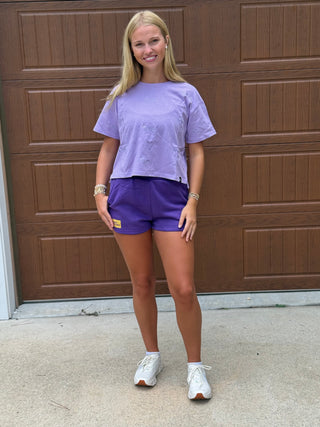 A woman stands outside wearing a light purple short-sleeve crop top with embroidered "Pirates" throughout and matching purple athletic shorts, with white sneakers.