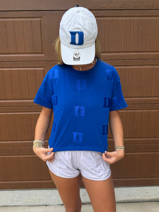 A woman wearing a bright royal blue short-sleeve t-shirt with embroidered "D" for Duke University paired with white shorts and a white baseball cap, standing in front of a brown garage door.