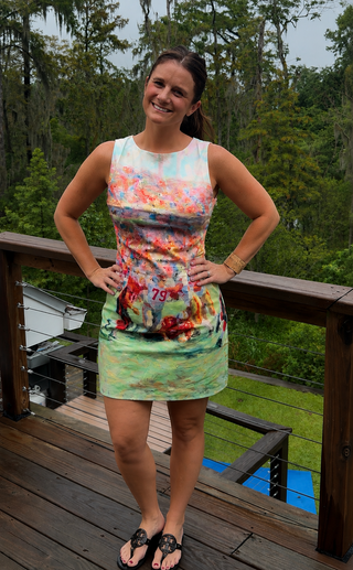 A woman stands on a wooden deck wearing a sleeveless, fitted mini dress with a colorful abstract football print. 