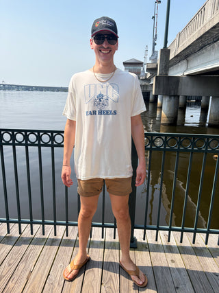 A man wearing a beige UNC tar heels graphic t-shirt, tan shorts, and a baseball cap stands on a wooden boardwalk railing.