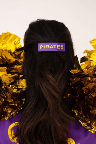 Person wearing a purple hair clip with 'PIRATES' text, surrounded by gold pom-poms on a white background