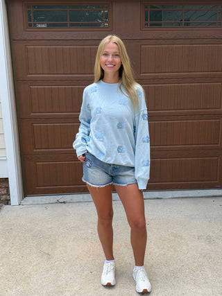 A young woman stands outside wearing a light blue long-sleeve sweatshirt with embroidered "UNC" throughout with denim shorts tar heel shorts, and white sneakers.