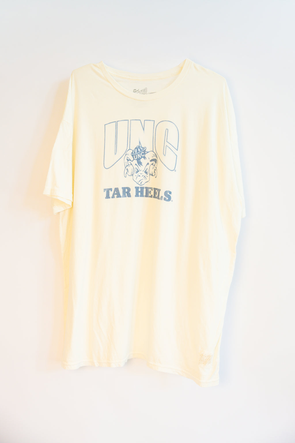 A pale beige short-sleeve t-shirt with a light blue graphic print on the front reading 'UNC TAR HEELS' with a ram mascot logo, displayed against a plain white background.