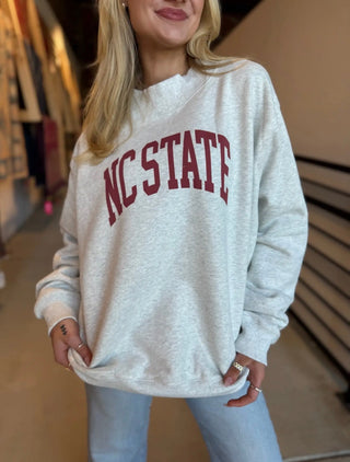 A woman wearing a light heather gray sweatshirt with 'NC STATE' printed in dark red arched letters across the chest, paired with light blue jeans in an indoor setting.