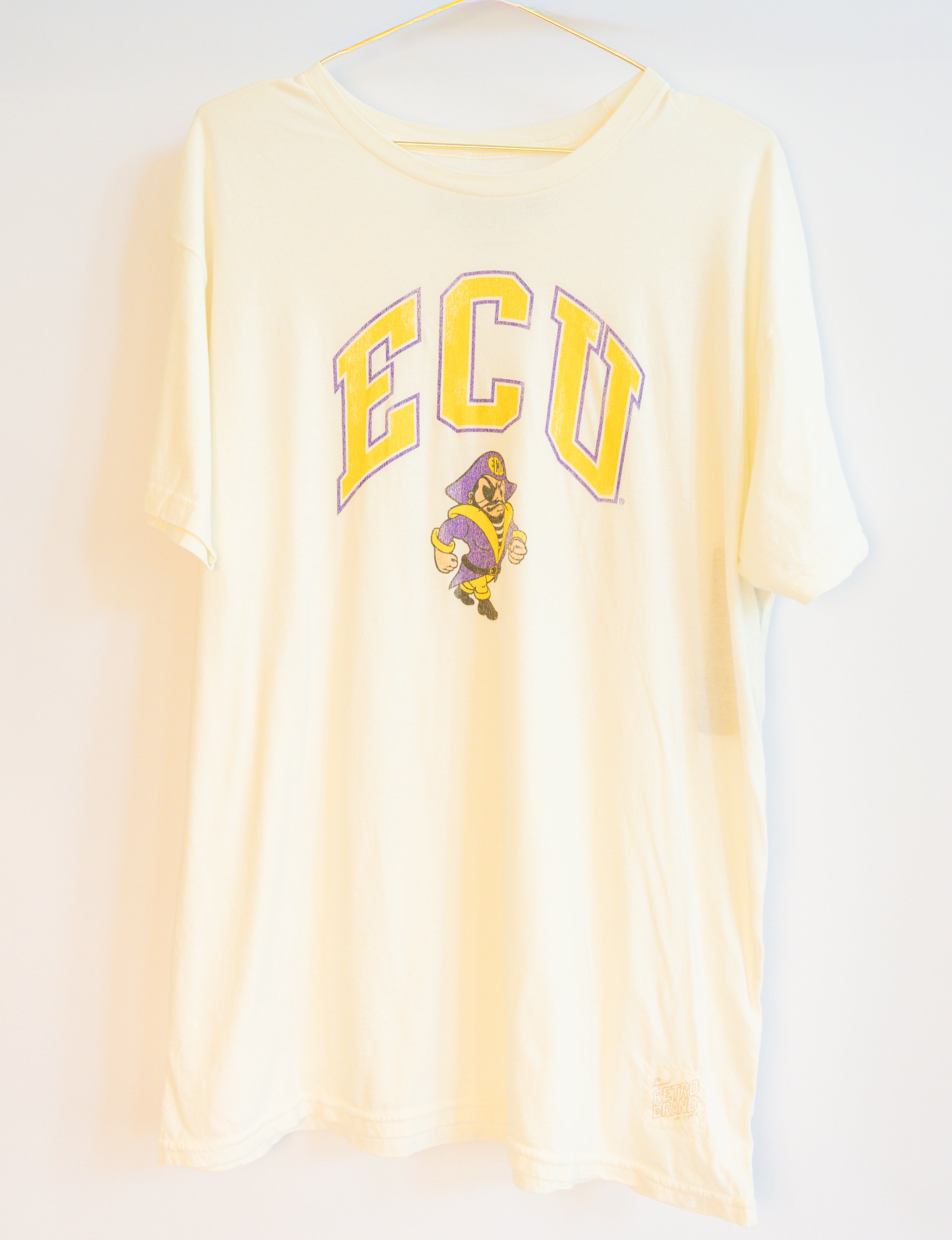 A cream-colored short-sleeve t-shirt hanging on a hanger against a white wall, featuring a yellow and purple 'ECU' graphic with the Texas Longhorns mascot on the front.