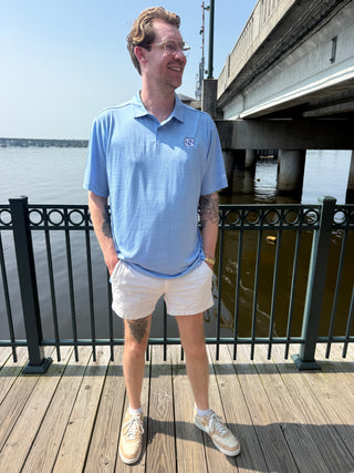 A man wearing a light blue UNC embroderied polo shirt, white shorts, and white sneakers stands on a wooden boardwalk next to a body of water with a bridge in the background.