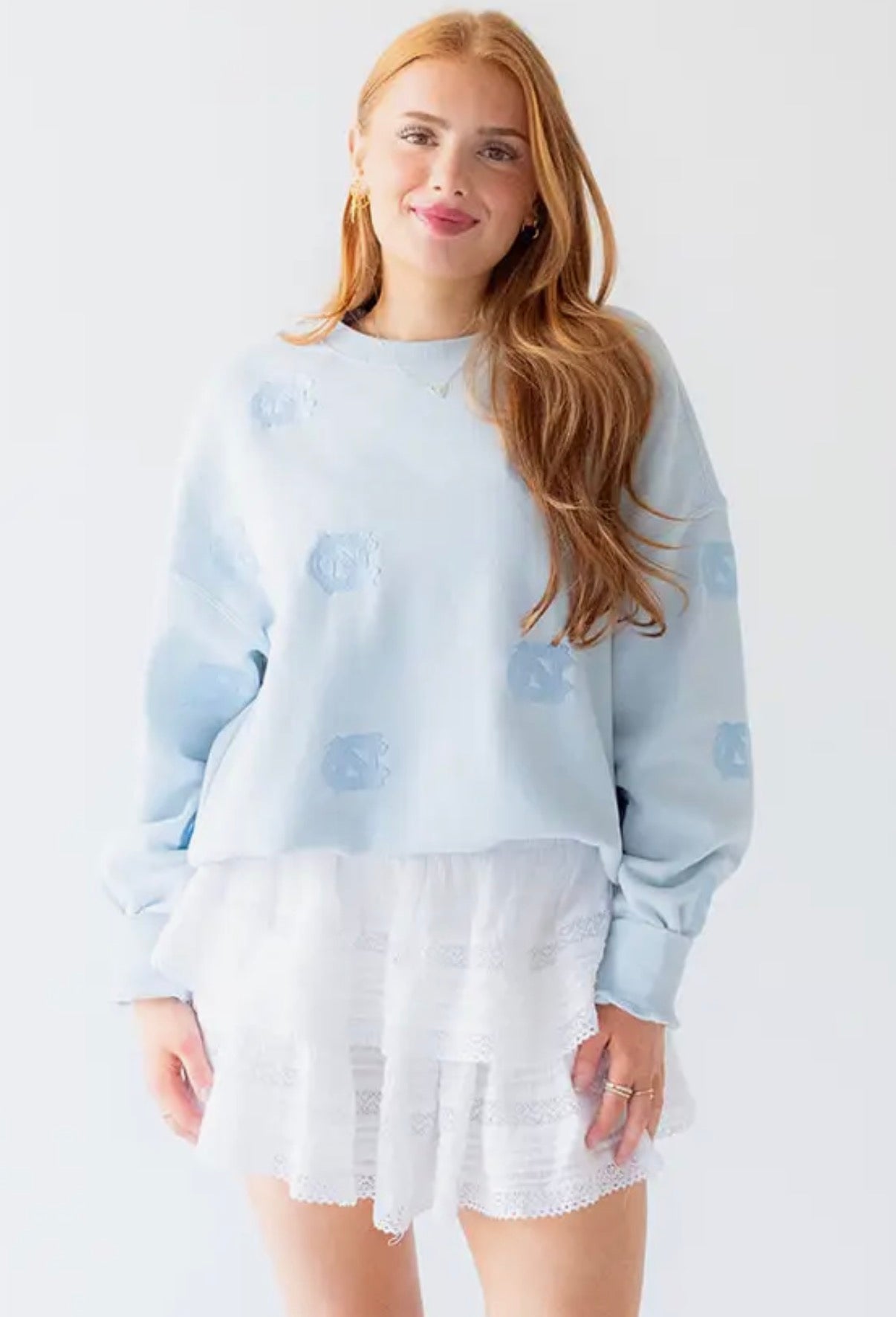 A woman modeling a light blue UNC crewneck sweatshirt with tonal embroidered details, paired with a white ruffled mini skirt, against a plain white studio background.
