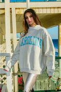 Woman wearing a reversible gray sweatshirt with 'CAROLINA' printed on it, standing on a beach deck.