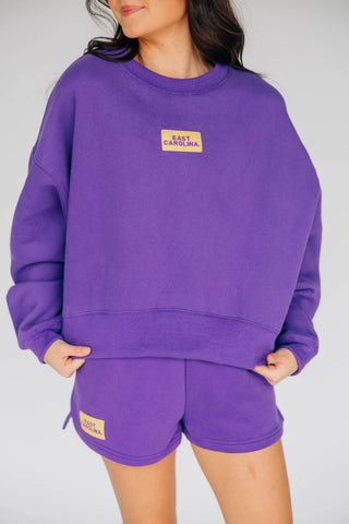 A person wearing a purple sweatshirt and matching shorts set, with a small tan rectangular patch on the chest and shorts that reads 'EAST CAROLINA'. The photo is cropped from the lower face to mid-thigh against a light gray studio background.
