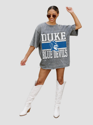 Woman wearing a gray rhinestone Duke Blue Devils t-shirt dress with white boots on a light gray background